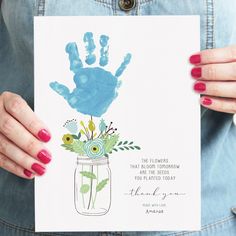 a woman holding up a card with a handprint on it and flowers in a mason jar