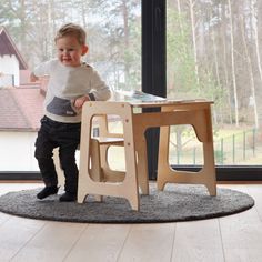 a toddler playing with a wooden table and chair in front of a large window