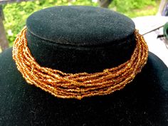 "Bead Choker with 12 strands of golden glass beads Small seed beads with a larger barrel bead about every 3\" Larger faceted bead fits in loop to close Adjustable size 13.5\" - 15.5\" 1.25\" wide Condition - very good" Gold Double Strand Beaded Necklaces With Tiny Beads, Gold Double Strand Beaded Necklace With Tiny Beads, Amber Beaded Necklaces With Gold Beads For Gifts, Gold Beaded Bracelets With Polished Beads For Festival, Gift Amber Beaded Necklaces With Gold Beads, Amber Beaded Necklace With Gold Beads For Gift, Gift Amber Beaded Necklace With Gold Beads, Gold Necklace With Colorful Beads For Festivals, Gold Beaded Bracelets With Colorful Beads For Festivals