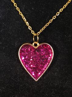 a heart shaped necklace with pink glitter on it sitting on a black surface, in the shape of a gold chain