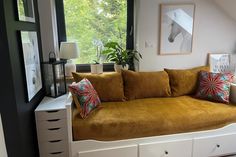 a couch with pillows on it in front of a window next to a dresser and lamp