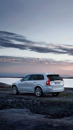 a white volvo suv parked on the side of a body of water at dusk with clouds in the sky