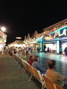 people are sitting on benches in front of the boardwalk at night time, with shops and restaurants lit up behind them