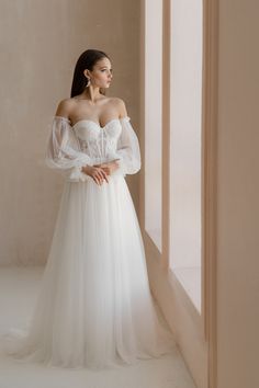 a woman in a white wedding dress standing by a window with her hands on her hips