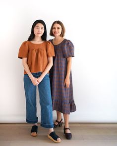 two young women standing next to each other in front of a white wall wearing brown tops and blue jeans