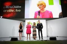 three women and one man standing on stage in front of a large screen with the words hello my children on it