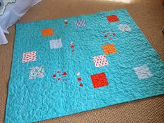 a blue quilt with orange and white squares on it sitting on the floor next to a bed