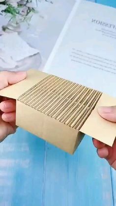 two hands holding a piece of brown paper over a blue wooden table with an open book in the background