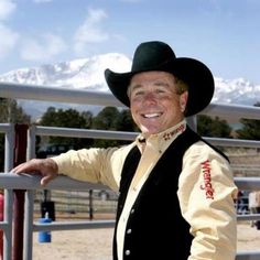 a man wearing a cowboy hat standing in front of a fence with mountains behind him