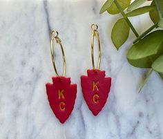 two red earrings with the letters kc and c on them sitting next to a plant