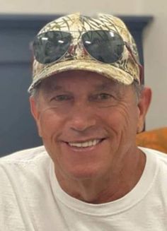 an older man wearing sunglasses and a camo hat smiles at the camera while sitting in a chair