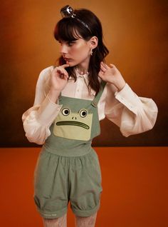 a woman in green overalls and white shirt with frog face on the front, holding her finger to her nose