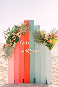 a colorful wooden sign with flowers on it sitting in the sand at the beach next to some palm leaves