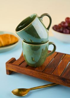 two green coffee cups sitting on top of a wooden tray next to grapes and croissants