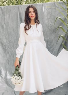 a woman in a white dress is holding a bouquet and standing next to a wall