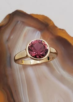 DEN2407 Pink tourmaline solitaire ring. Elegant bezel set ring designed for comfort and style set with Maine Pink Tourmaline.  Ring mounting is low profile 14k yellow gold.  Round brilliant cut pink tourmaline is 2.20cts, approximately 8.5mm in diameter. This stone is from the 1972 Dunton Quarry Newry, ME discovery. Currently the ring is size 5.5 but we will resize at no extra charge if needed. Please specify ring size at checkout. Tension Ring Settings, Pink Stone Rings Gold, Pink Tourmaline Cabochon Ring, Modern Tourmaline Rings With Bezel Setting, Tourmaline Birthstone Ring With Bezel Setting, Classic Pink Rings With Bezel Setting, Classic Pink Bezel Set Ring, Classic Pink Ruby Ring With Bezel Setting, Pink Tourmaline Jewelry