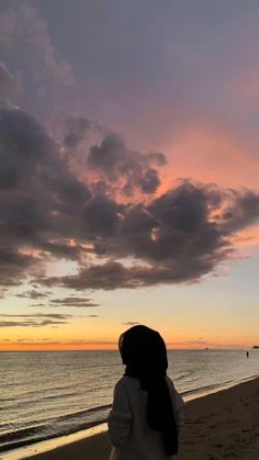 a woman standing on top of a sandy beach next to the ocean at sunset with clouds in the sky