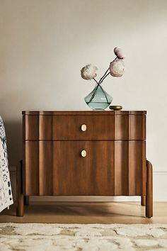 a wooden cabinet with two flowers on top and a bed in the background, along with a white rug