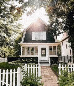 a white picket fence surrounds the front yard of a small black and white house with sun shining through the windows