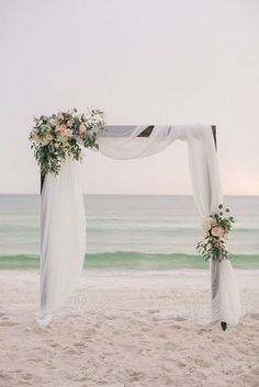 an arch decorated with flowers and greenery is on the sand at the beach near the ocean