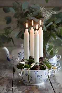 four white candles are sitting in a bowl with moss and pine cones on the table