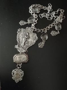 The central pendant in this assemblage is an art nouveau plaque in plated silver that most likely adorned the back of a mirror. It supports an antique Victorian aesthetic movement sterling brooch featuring a flying swallow. From the brooch is a hallmarked British sterling Victorian era watch fob with a gilded initialed cartouche. Accompanying pendants are rhodium plated bezel set free form faceted crystals and Victorian inspired filigree charms. All elements are attached to silver plated book chain and the necklace is finished with larger links and a hook so you can adjust the length. Silver Medallion Art Deco Jewelry, Silver Art Deco Medallion Necklace, Elegant Silver Medallion Brooch, Art Deco Silver Jewelry With Vintage Charm, Silver Art Deco Jewelry With Vintage Charm, Victorian Silver Necklace With Antique Finish, Silver Art Nouveau Brooch, Antique Silver Pendant Brooches, Silver Medallion Brooches For Collectors