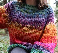 a woman wearing a multicolored sweater sitting in the grass with her hands on her hips