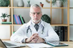 a doctor sitting at a desk in front of a laptop computer and holding his hands together