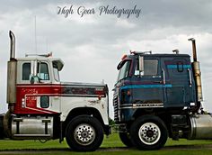 two semi trucks parked on the grass near each other with cloudy skies in the background