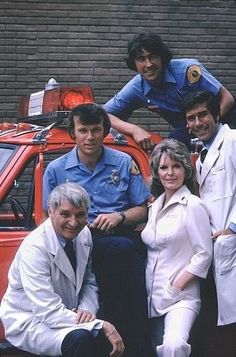 four people posing in front of a red car with police officers on top and behind them