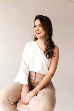 a woman sitting on top of a stool wearing beige pants and a white shirt with her hands in her pockets