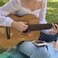 a woman sitting on the grass playing an acoustic guitar with her cell phone in her lap