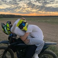 two people sitting on top of a motorcycle in the middle of a dirt field at sunset