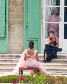 two women sitting on a bench in front of green shuttered windows, one taking a photo