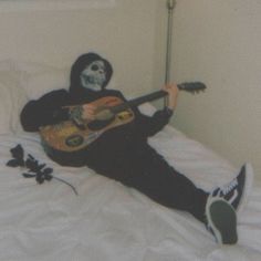 a person in a skeleton mask playing guitar on a bed with white sheets and pillows