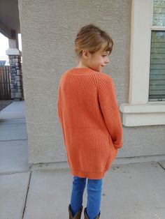 a young boy standing on the sidewalk wearing an orange sweater and blue jeans with brown boots