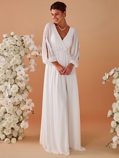 a woman standing in front of white flowers wearing a long sleeved wedding dress with an open back