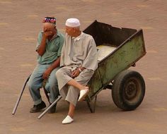 two older men sitting on top of a green wheelbarrow next to each other