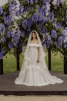 a woman wearing a wedding dress and veil standing in front of purple flowers on a tree