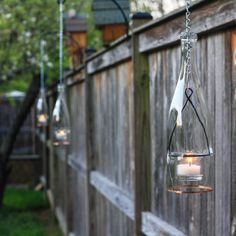 three hanging glass jars filled with candles on a fenced in backyard area that has a wooden fence and green grass