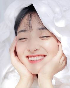 a woman with her eyes closed under a white blanket smiling and looking at the camera