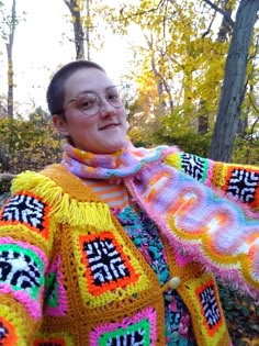 a woman wearing glasses and a colorful crochet sweater in the woods with her arms outstretched