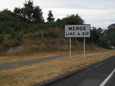 a sign that says merge like a zip on the side of a road with trees in the background