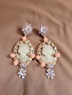 The earrings have a light brown Victorian female carved portrait.  3.5 mm enameled pink roses and golden leaves are on the border of an oval frame.  The post has a flower shaped created diamond with a one carat round center and .5 carat diamond petals.  The bottom has a one carat diamond flower.  18k gold plated. Elegant Cameo Jewelry For Wedding, Cameo Drop Earrings As Gift, Cameo Drop Earrings Jewelry Gift, Gift Cameo Drop Earrings Jewelry, Luxury Oval Pink Earrings, Luxury Pink Oval Earrings, Elegant Cameo Earrings, Elegant Cameo Jewelry, Elegant Cameo Jewelry For Formal Occasions