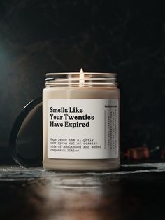 a candle sitting on top of a wooden table next to a black mug with the words smell like your twenties have expired