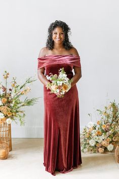 a woman standing in front of flowers wearing a red velvet dress with off the shoulder sleeves