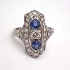 Art Deco 14k (.585) white gold long ring, decorated with Diamonds and Sapphire stones. There are 22 single cut Diamonds, approx. 1.55 mm, the center stone is Old Mine Cut 4.67 x 4.2 x 3.25 mm, color K, SI1 clarity. 2 natural Sapphires are 4.27 mm in diameter. This stately ring is a size 5, it is 22 mm long, weighing 2.7 grams. EA4815 Multi-stone Round Sapphire Ring In Platinum, Art Deco Platinum Sapphire Ring With 17 Jewels, Art Deco Platinum Sapphire Ring, Art Deco Multi-stone Diamond Ring, Round Cut Diamond Ring In Platinum, Platinum Diamond Ring With Round Cut Gemstone, Art Deco Diamond White Ring With 17 Jewels, Diamond White Marquise Multi-stone Ring, Diamond White Multi-stone Marquise Rings