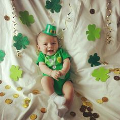 a baby wearing a green shirt and hat sitting in front of shamrocks on a white sheet