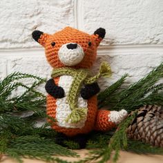 a crocheted stuffed animal with a scarf around it's neck sitting next to pine branches