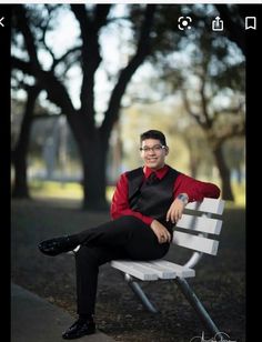 a man in a red shirt and black pants sitting on a white bench with trees behind him